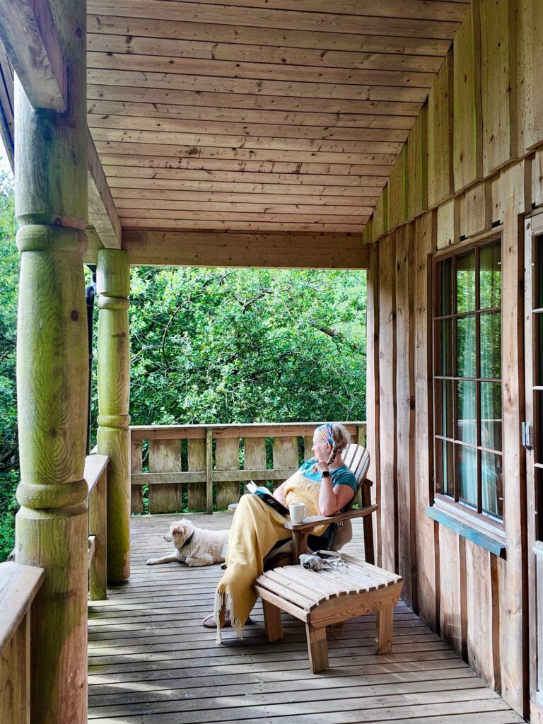 Cookie a white fluffy dog sat next to a wooden arm chair with blankets, a book and a tea on it